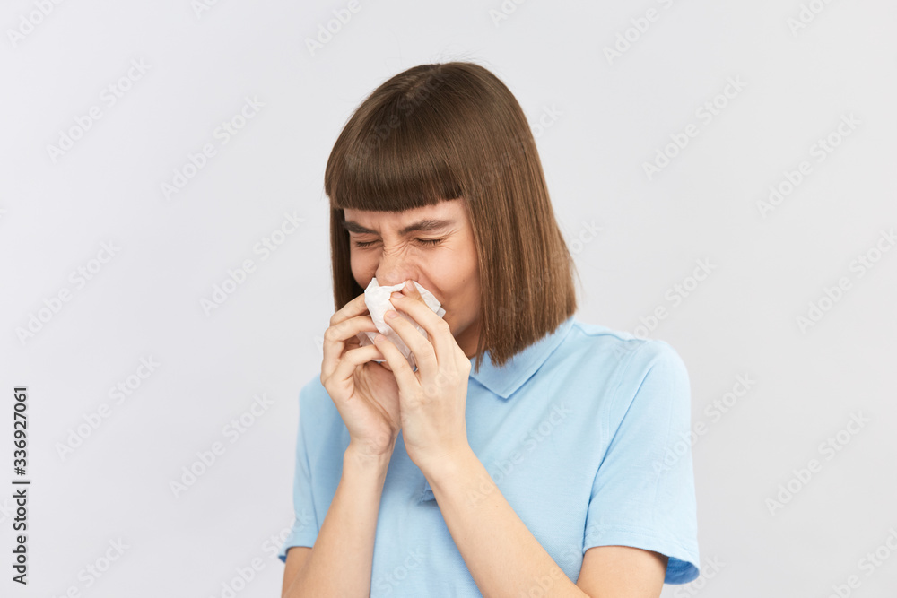 female with dark hair and in blue shirt blows nose with eyes closed on grey background female with dark hair and in blue shirt blows nose with eyes closed on grey background