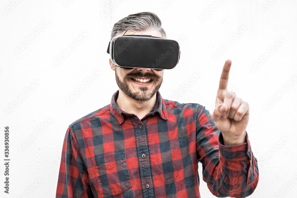 Fototapeta premium Content man in VR headset pointing with finger. Front view of cheerful bearded man in checkered shirt using virtual reality headset on grey background. Technology concept