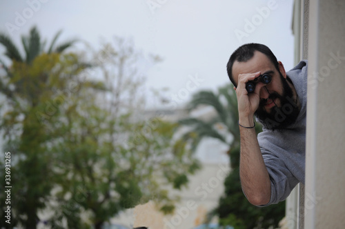 bald man with long beard leaning out from building window looking through binoculars. Picture with copy space of man using binoculars to spend time on times of quarantine