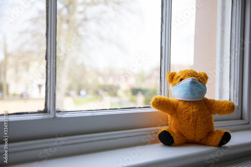 A teddy bear wearing a protective medical mask sitting by a window in isolation