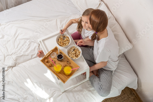 Happy mom and daughter have healthy breakfast on bed in a light bedroom on a sunny morning. Orange juice and oatmeal porridge with nuts and raisins. Healthy food concept. Top view