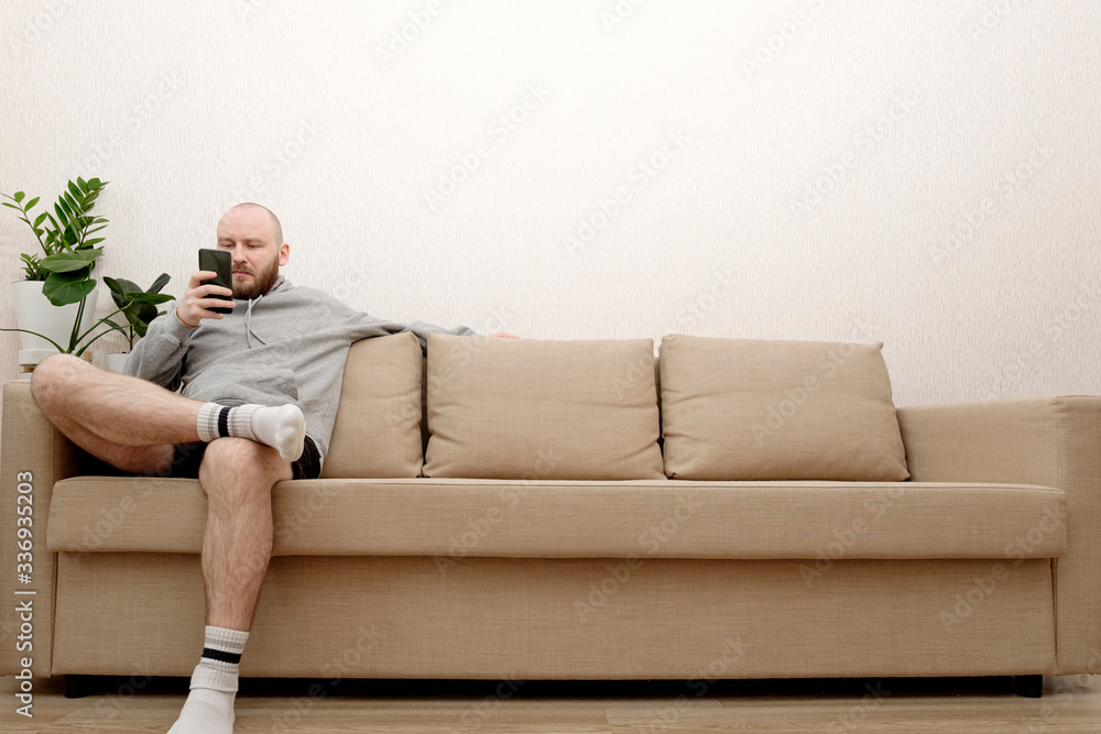 A young man with a beard in home clothes is sitting on a sofa and is using a smartphone. Smartphone - salvation from loneliness during self-isolation. The guy stayed home so as not to get a coronoviru
