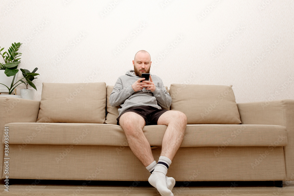 A young man with a beard in home clothes is sitting on a sofa and is using a smartphone. Smartphone - salvation from loneliness during self-isolation. The guy stayed home so as not to get a coronoviru