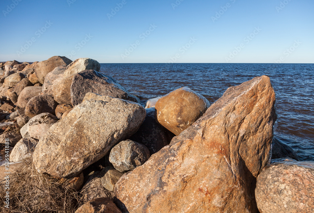 Huge boulders covered with ice against the blue water of Lake Ladoga on a sunny day in early spring