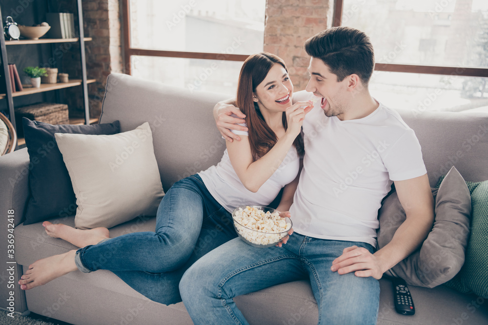Portrait of nice attractive lovely cheerful cheery funny couple sitting on divan watching TV having fun eating corn quarantine at modern industrial loft style brick interior living-room flat