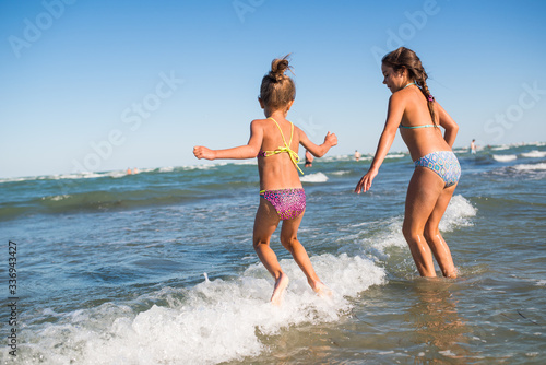 Two funny little girls jump in the noisy sea waves and enjoy the long-awaited vacation on a sunny warm summer day. Concept of sea vacation and travel with children © YouraPechkin