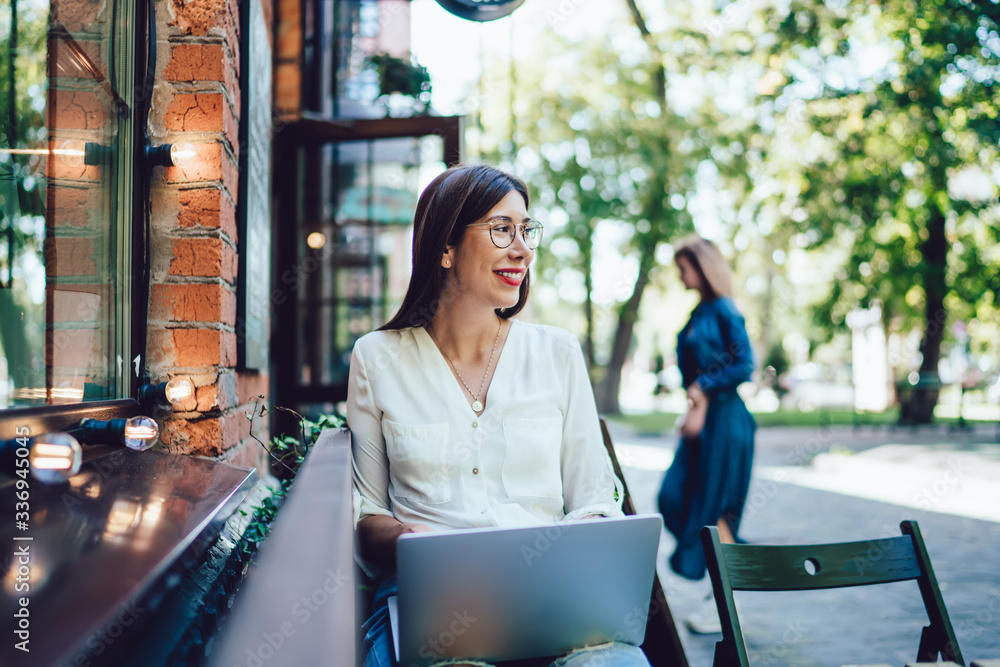 Positive hipster girl with cute smile on face looking away while waiting distance colleague in coworking terrace for freelancers, happy female with in optical glasses using laptop computer