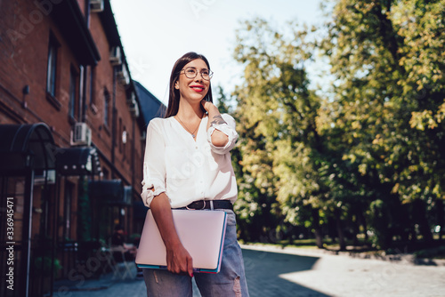 Cheerful charming woman in classic eyewear holding laptop for e learning and smiling at city urban setting, young millennial female student trendy dressed looking away and feeling good outdoors