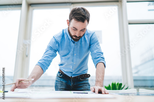 Adult man intently working with blueprint while standing at desk in open space office