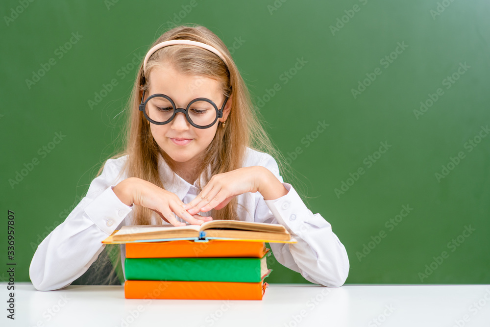 Young girl wearing funny eyeglasses read book near empty green chalkboard. Empty space for text Young girl wearing funny eyeglasses read book near empty green chalkboard. Empty space for text