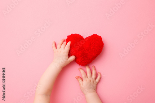 Baby girl hands touching bright red heart. Light pink background. Pastel color. Love concept. Point of view shot. Closeup. Top down view.