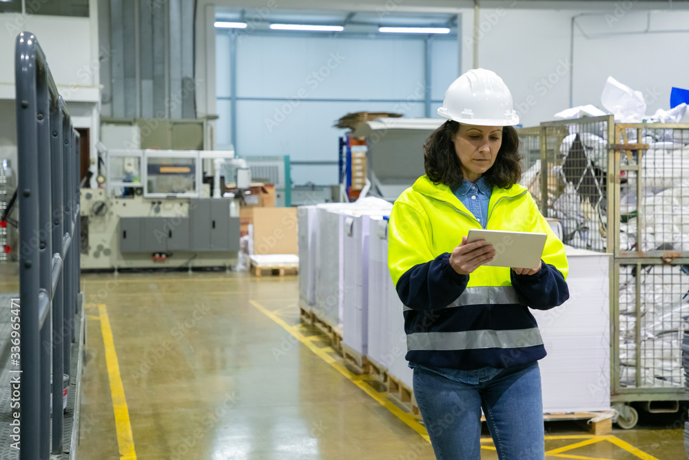 Focused female engineer with tablet computer monitoring equipment onsite. Middle aged woman in hardhat and uniform walking on plant floor. Machinery or electronic control concept Focused female engineer with tablet computer monitoring equipment onsite. Middle aged woman in hardhat and uniform walking on plant floor. Machinery or electronic control concept
