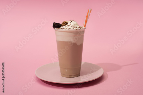 Disposable cup of chocolate milkshake with drinking straw on plate on pink background