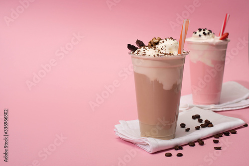 Selective focus of disposable cups of chocolate and strawberry milkshakes with coffee grains on napkins on pink background