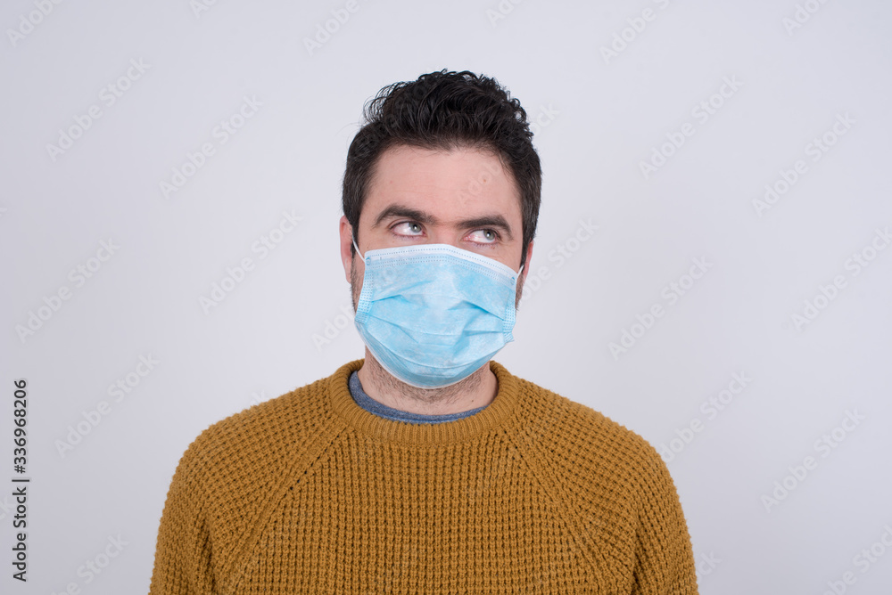 Gloomy, wondering young man face looking up lwearing medical face mask, being upset thinking about something important, keeps hands down, man standing indoors. Gloomy, wondering young man face looking up lwearing medical face mask, being upset thinking about something important, keeps hands down, man standing indoors.