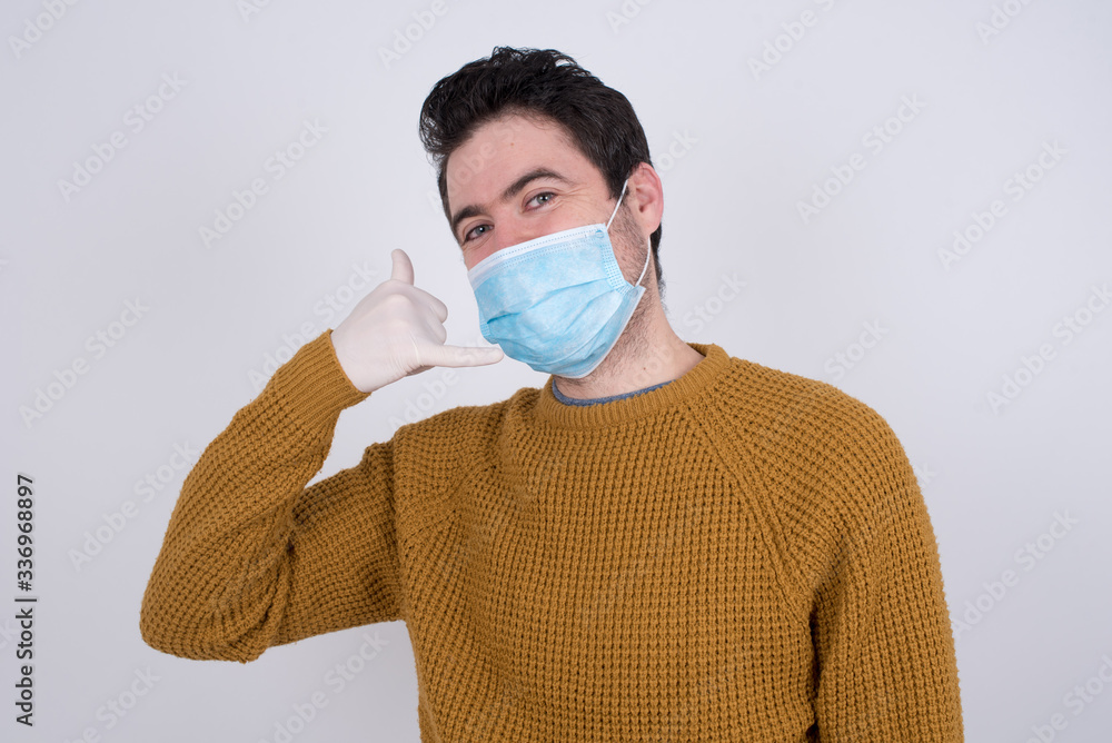 Attractive young male wearing medical face mask imitates telephone conversation, keeps hand near ear as if holding mobile phone, has confident facial expression, isolated outdoors. Call me! Attractive young male wearing medical face mask imitates telephone conversation, keeps hand near ear as if holding mobile phone, has confident facial expression, isolated outdoors. Call me!