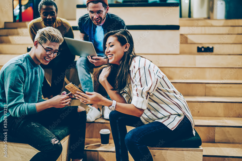 Happy male and female teen students share multimedia files online via smartphones during meeting in college, skilled positive multiracial crew of employees using technologies for research together