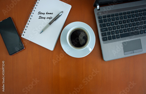 A desk with a cup of black coffee, laptop and a notepad with letters Stay home, stay safe on it with a pen during COVID pandemic