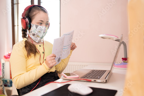 Young girl in antiviral mask studying homework during online lesson at home, social distance during quarantine, self-isolation. Online education concept. Home schooler. Student shows notes to teacher.