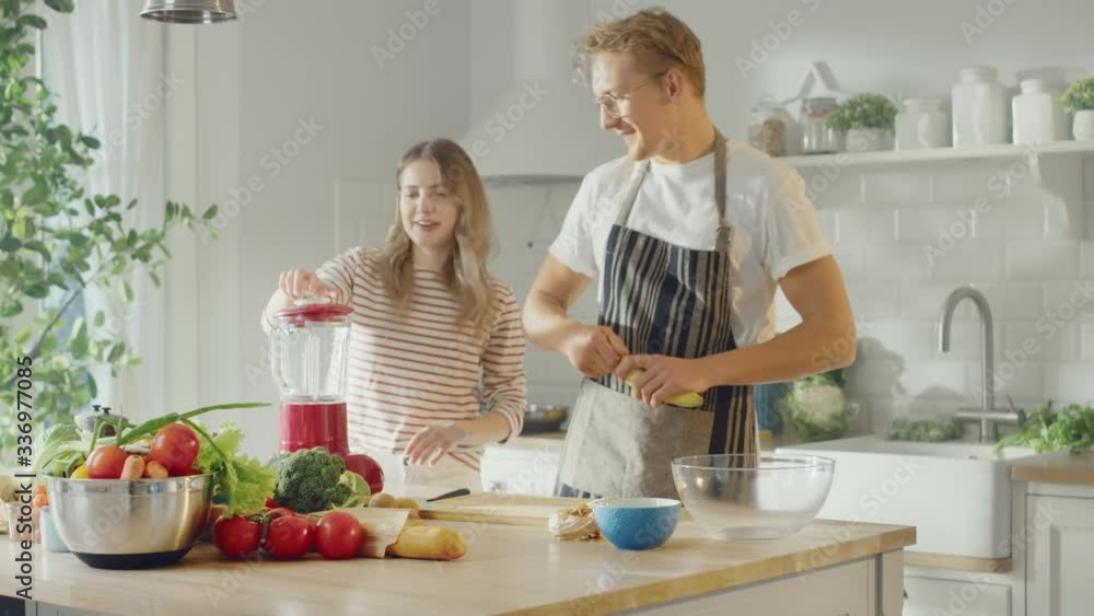 Handsome Young Man in Glasses Wearing Apron and Beautiful Girl are Making A Smoothie in the Kitchen. Happy Couple are Preparing Healthy Organic Beverage. Male and Female at Home on a Sunny Day. Handsome Young Man in Glasses Wearing Apron and Beautiful Girl are Making A Smoothie in the Kitchen. Happy Couple are Preparing Healthy Organic Beverage. Male and Female at Home on a Sunny Day.
