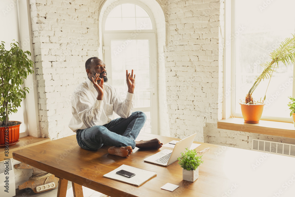 Young african-american man doing yoga at home while being quarantine and freelance online working. Remote, isolated or alone at office. Concept of healthy lifestyle, wellness, activity, movement. Young african-american man doing yoga at home while being quarantine and freelance online working. Remote, isolated or alone at office. Concept of healthy lifestyle, wellness, activity, movement.