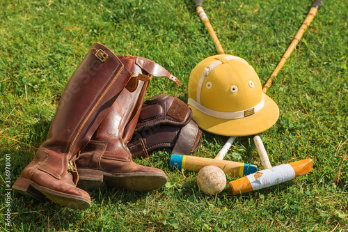 Equipment for a player in horse polo lies on a green lawn. Leather riding boots, protective helmet on the head, knuckles, ball and crossed mallets as a sports game concept. Top view