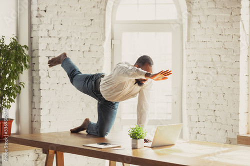 Young african-american man doing yoga at home while being quarantine and freelance online working. Remote, isolated or alone at office. Concept of healthy lifestyle, wellness, activity, movement. photo