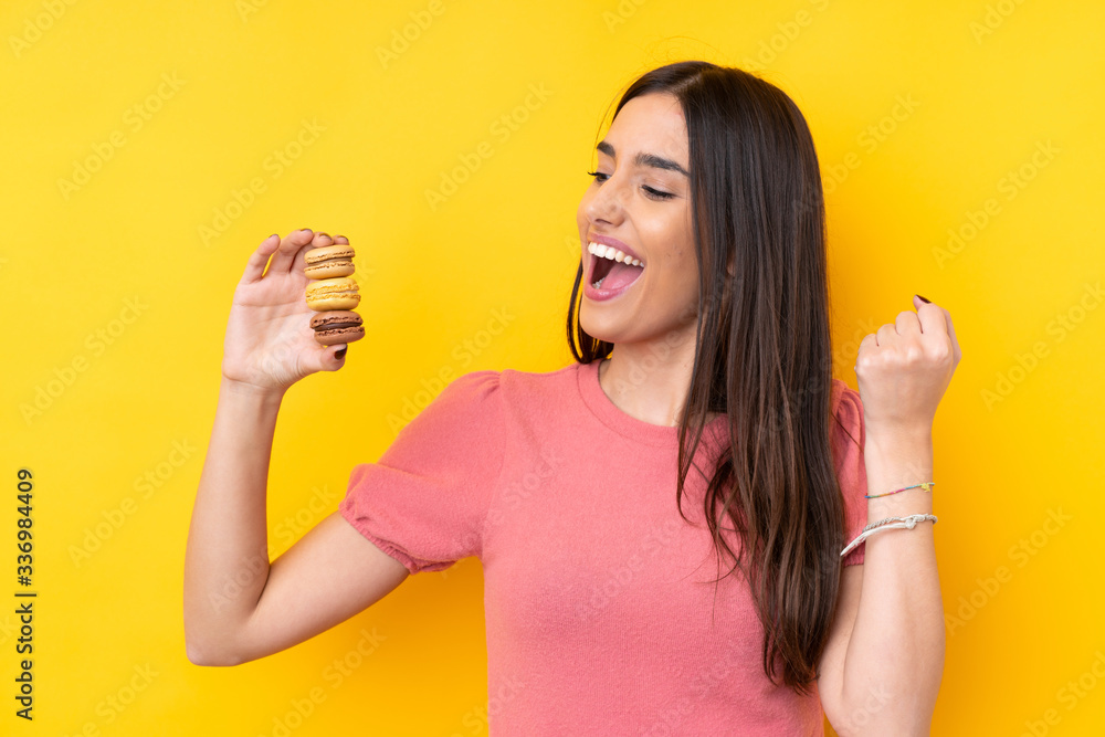 Young brunette woman over isolated yellow background holding colorful French macarons and celebrating a victory Young brunette woman over isolated yellow background holding colorful French macarons and celebrating a victory