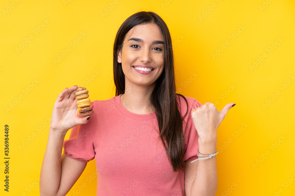 Young brunette woman over isolated yellow background holding colorful French macarons and pointing side Young brunette woman over isolated yellow background holding colorful French macarons and pointing side