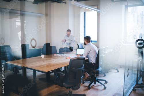 Young prosperous male designers having meeting in coworking office discussing project ideas satisfied with successful collaboration, smiling 20s businessmen talking to each other share opinions