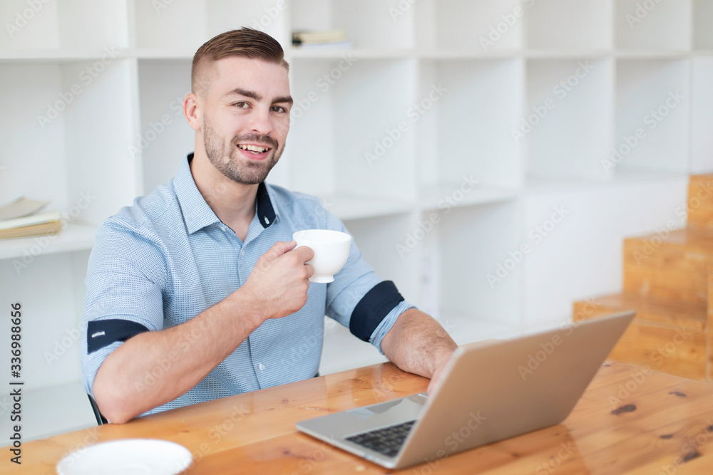 Young man drinking coffee while working with laptop. Hand man drinking coffee in office while typing on laptop in a cafe Young man drinking coffee while working with laptop. Hand man drinking coffee in office while typing on laptop in a cafe