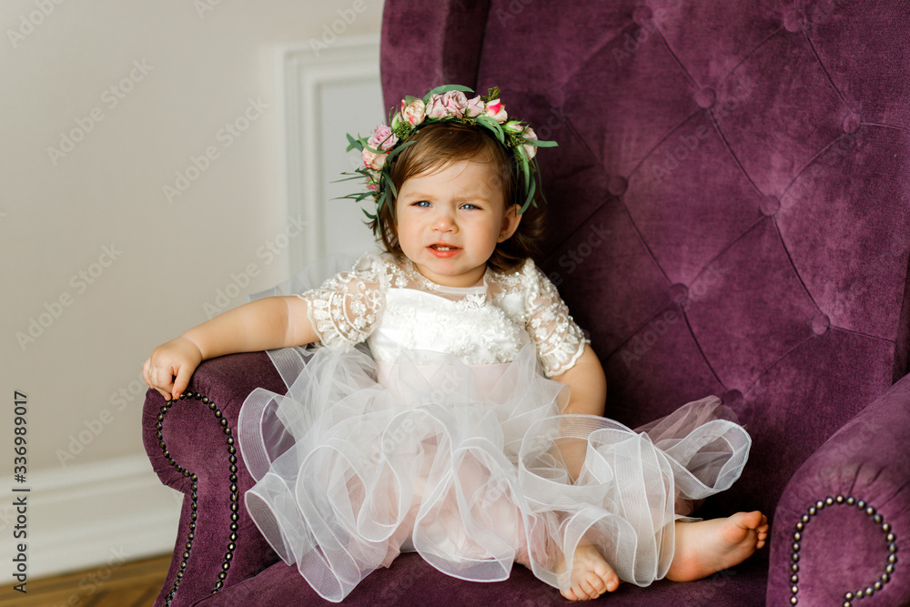 Sweet little girl in a pink dress sits on a soft chair. Crying baby ...