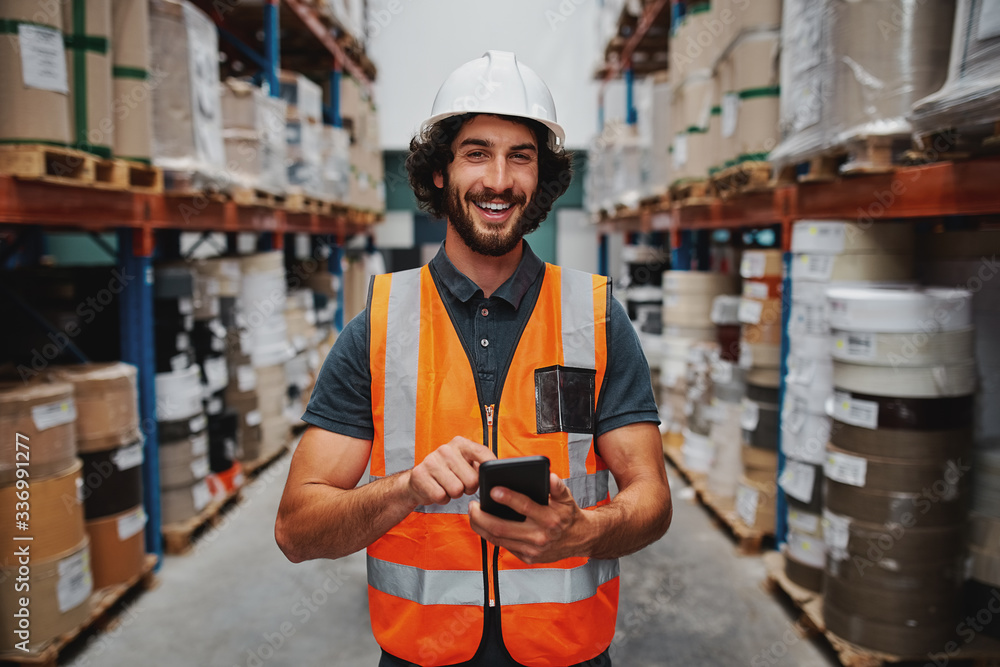 Young smiling male worker in warehouse wearing safety helmet and vest  standing between shelf with goods using cellphone Stock Photo | Adobe Stock