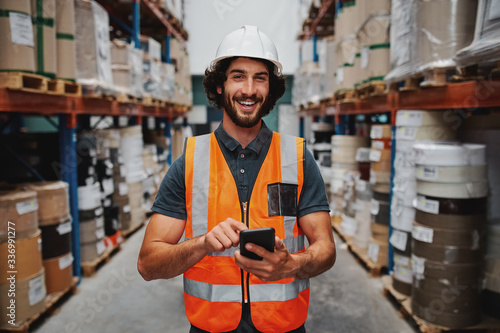 Young smiling male worker in warehouse wearing safety helmet and vest standing between shelf with goods using cellphone photo