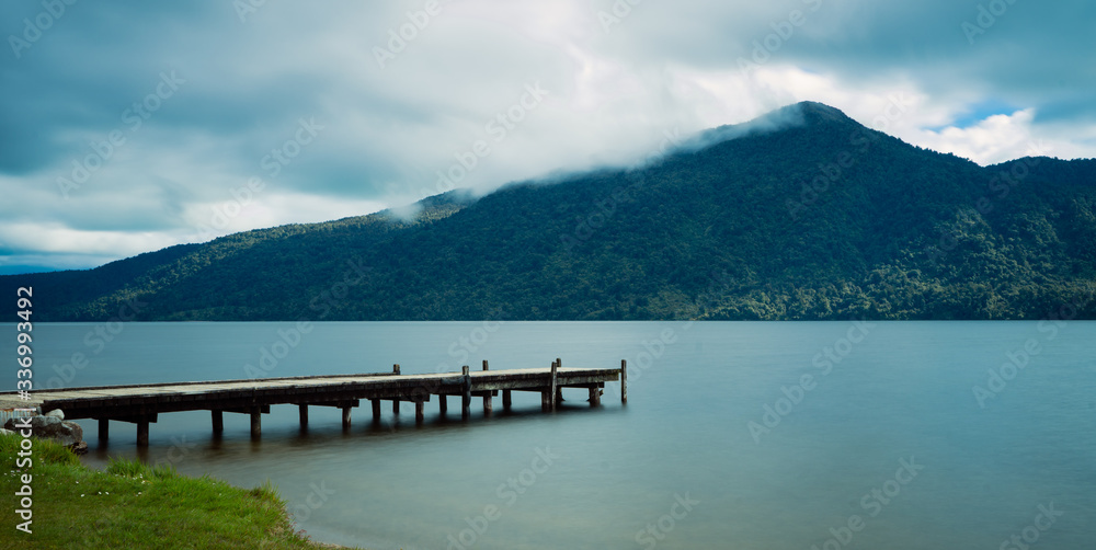 Beautiful panorama of a pier on Kaniere Lake with the green mountain in the background on a cloudy day, New Zealand