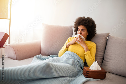 Sick young woman sitting on sofa blowing her nose at home in the sitting room. Photo of sneezing woman in paper tissue. Picture showing woman sneezing on tissue on couch in the living-room © Dragana Gordic