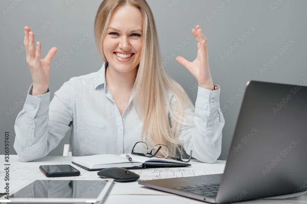Attractive happy business girl, being in a high spirit, smiles and raises her hands to the top with happiness, sitting behind her workplace. Attractive happy business girl, being in a high spirit, smiles and raises her hands to the top with happiness, sitting behind her workplace.
