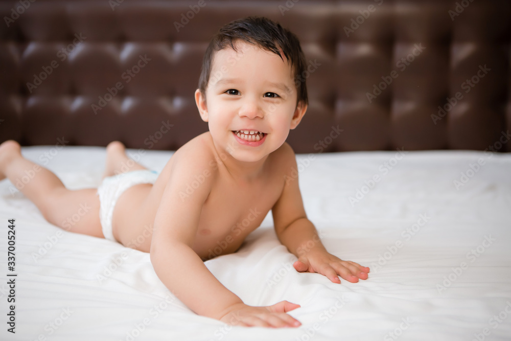 Cute toddler boy in diapers lies on bed with white sheet
