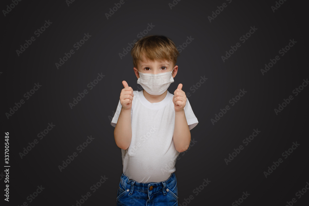 Little boy on a gray studio background in a medical mask holds thumbs up Little boy on a gray studio background in a medical mask holds thumbs up