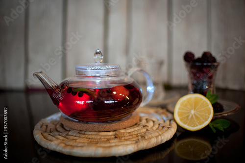  red tea with Basil and berries in a transparent round teapot on a wooden stand, black table wooden background