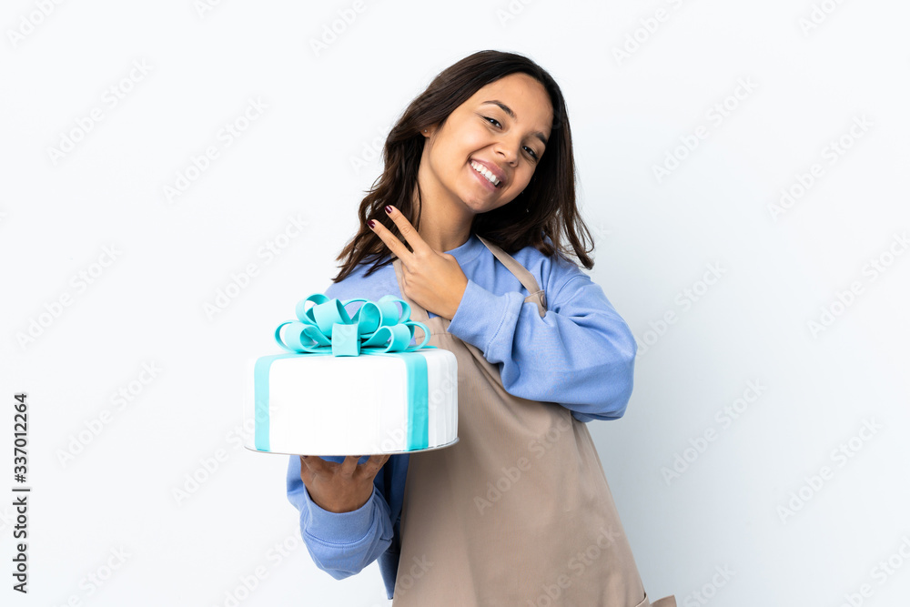 Pastry chef holding a big cake over isolated white background smiling and showing victory sign Pastry chef holding a big cake over isolated white background smiling and showing victory sign