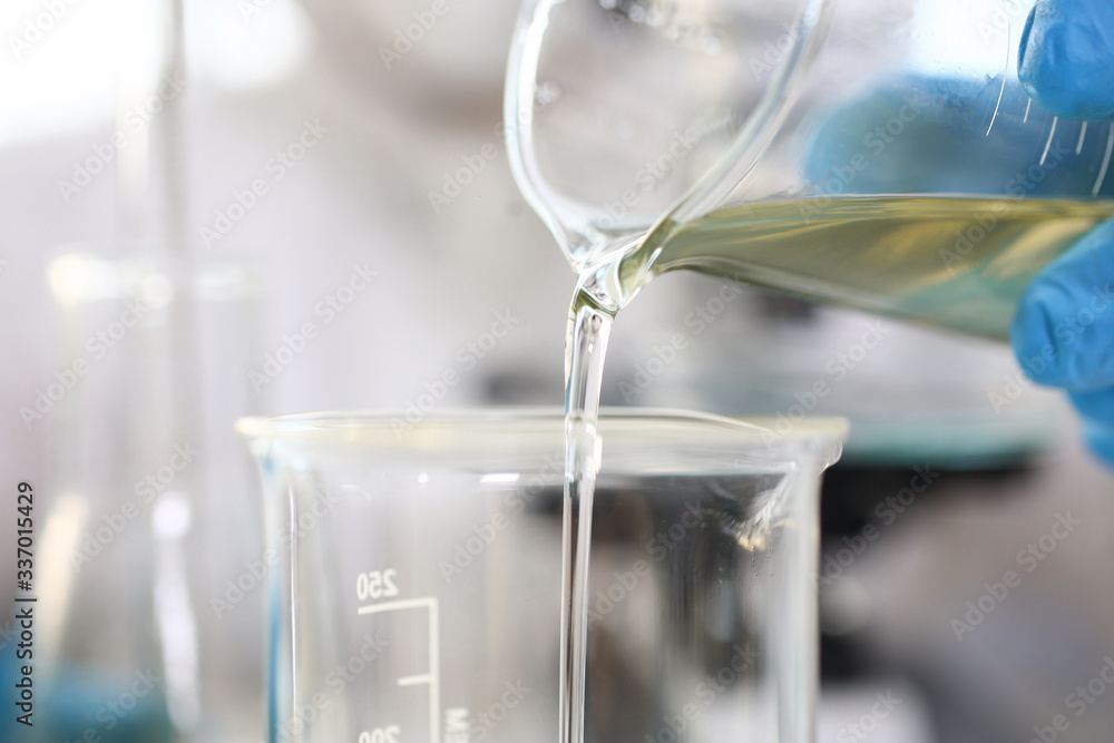 Scientist arm wearing blue protective gloves pouring yellow liquid at laboratory flask Scientist arm wearing blue protective gloves pouring yellow liquid at laboratory flask