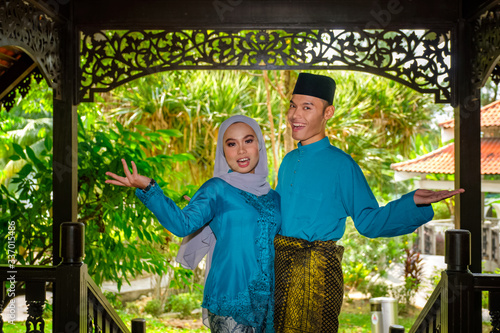 A portrait of young couple of malay muslim in traditional costume during Aidilfitri celebration showing welcome gesture by traditional wooden house photo