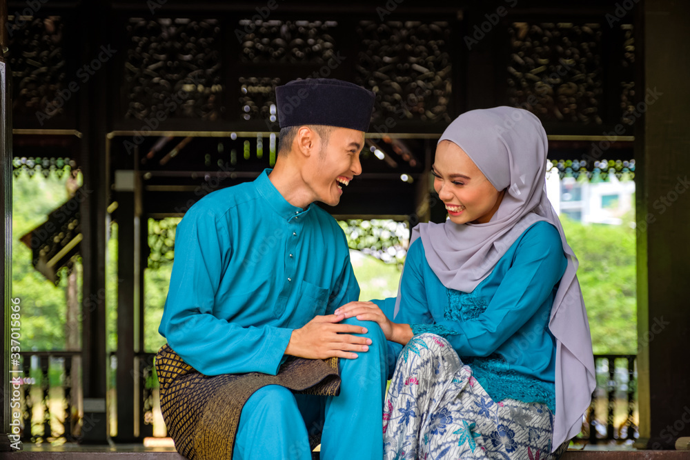 Young couple of malay muslim in traditional costume having happy conversation during Aidilfitri celebration at traditional wooden house.