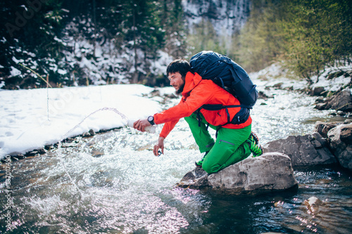 Fototapeta Naklejka Na Ścianę i Meble -  Young male tourist fillings water into the bottle from a river in the mountains..Beautiful winter landscape with snow covered banks and trees  on background. Climbing, trekking, active life concept.