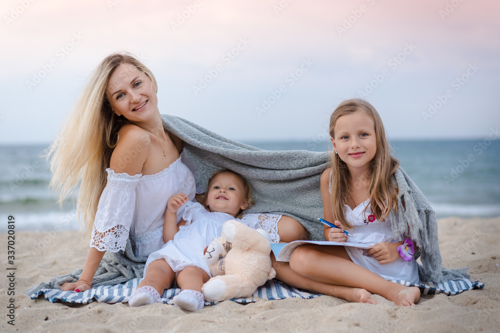 Mom with daughters blondes in white dresses laugh, hug, reading a book, hiding behind a knitted gray plaid, and sit near the blue sea on the beach at sunset. Mom with daughters blondes in white dresses laugh, hug, reading a book, hiding behind a knitted gray plaid, and sit near the blue sea on the beach at sunset.