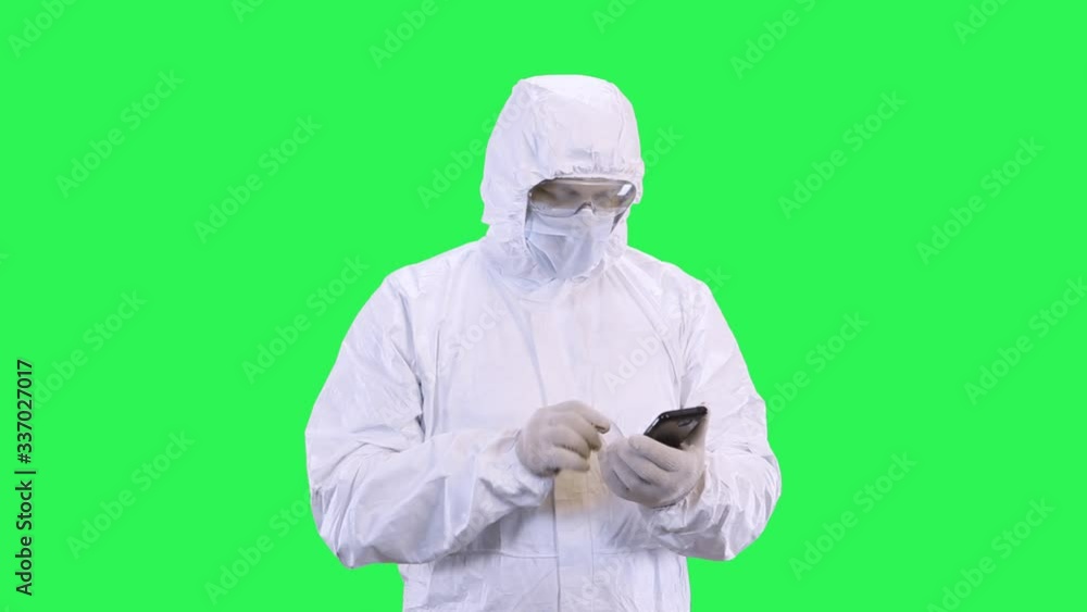 Masked man in protective suit surfing the phone while standing against green background Masked man in protective suit surfing the phone while standing against green background