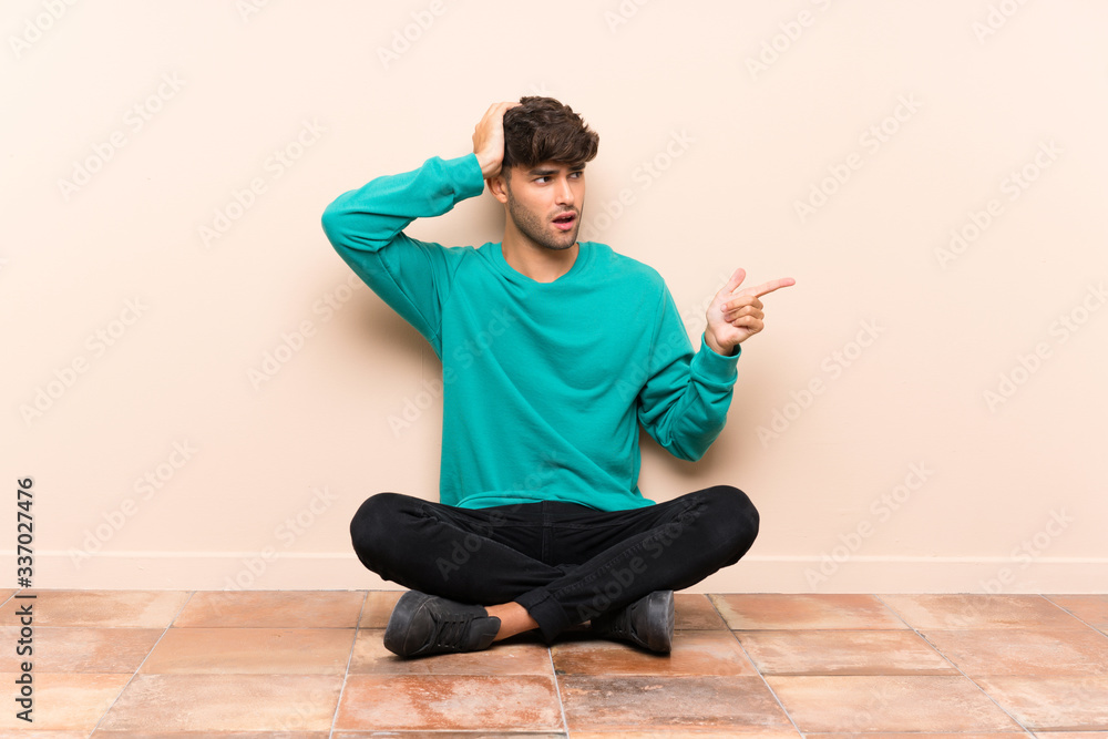 Young handsome man sitting on the floor surprised and pointing finger to the side Young handsome man sitting on the floor surprised and pointing finger to the side