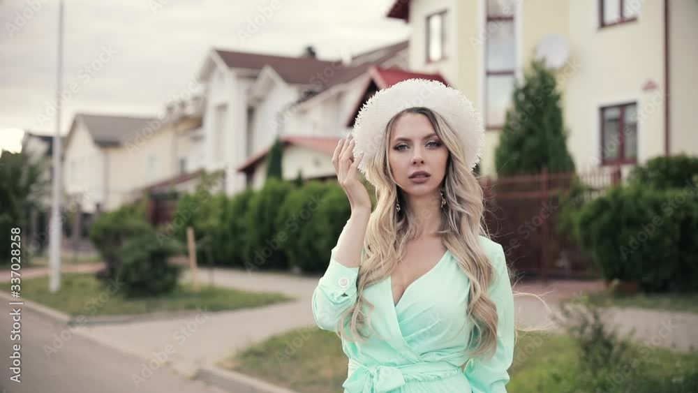 Young caucasian female with long blonde hair, white hat on her head in long mint dress and shoes poses for the camera Young caucasian female with long blonde hair, white hat on her head in long mint dress and shoes poses for the camera