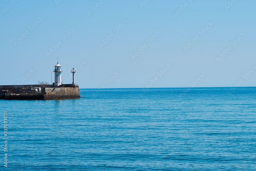 Lighthouse on the background of the sea and the sky, the sea is full calm and clear clear sky, spring. Lighthouse, sea and pier.